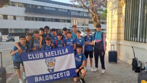 Group of Melilla Baloncesto junior players in blue uniforms posing with a banner outside Estación Marítima, luggage nearby.
