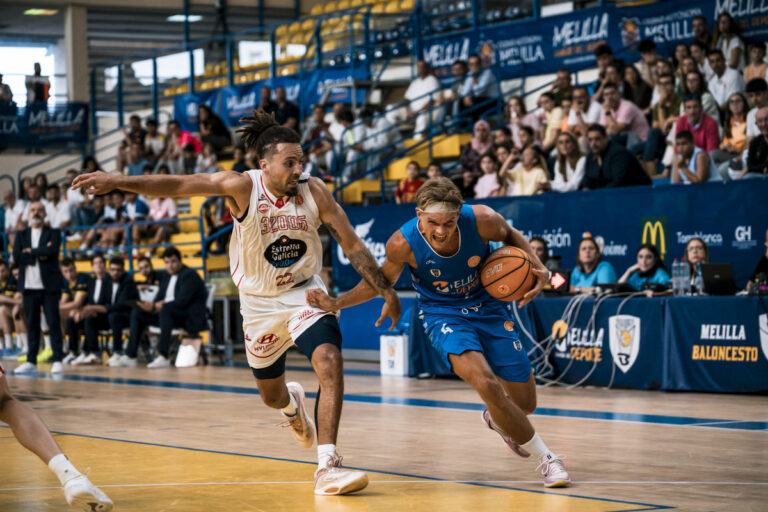 Basketball player in blue drives with the ball as a defender in white reaches in, with a crowded arena in the background.
