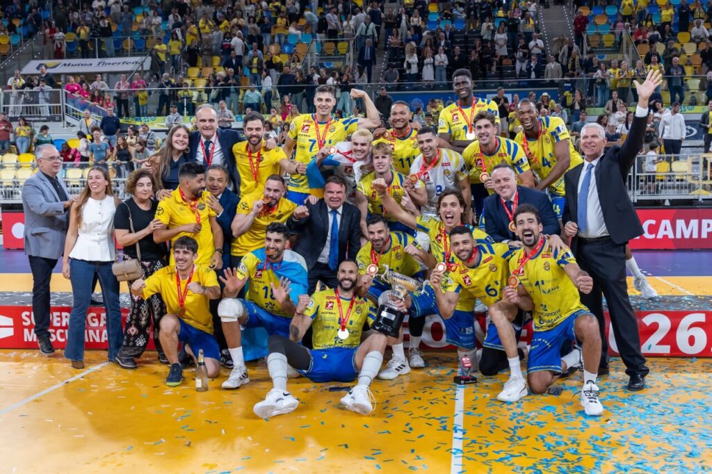 Volleyball team in yellow jerseys posing with trophy on court after victory, confetti around and fans cheering in the stands.