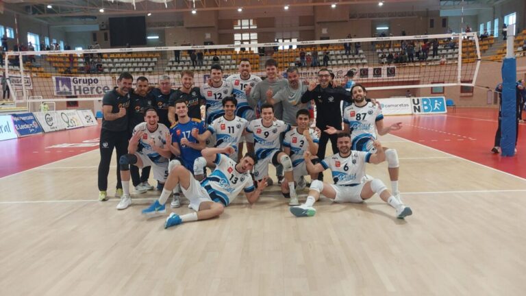Group of volleyball players posing on a court after a match, in white and blue uniforms, celebrating together with coaches in the back left.