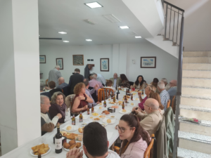 People seated around a long banquet table enjoying a meal with bread, wine, and plates of food in a bright room with framed art on the walls.