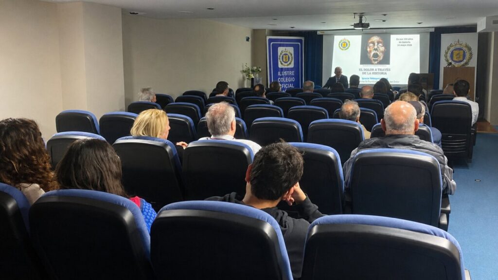 Audience seated in a small auditorium watching a presentation on a screen at the front of the room.
