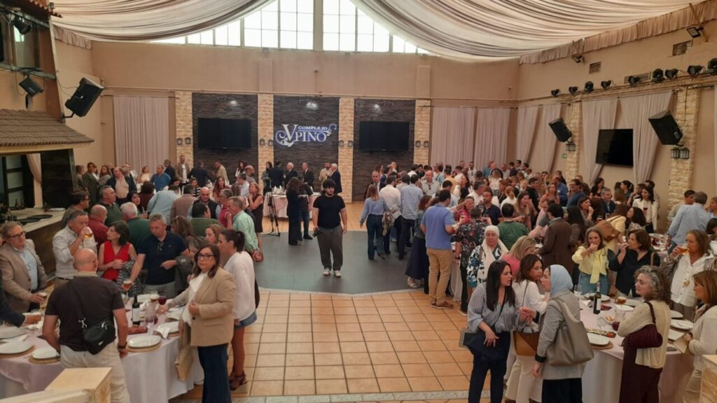 Busy banquet hall full of guests mingling around round tables with plates of food; stage back with screens and a banner above the stone backdrop.