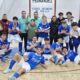 Group of young male futsal players in blue uniforms posing with coaches after a match in an indoor gym. Captioned banner behind reads team name.