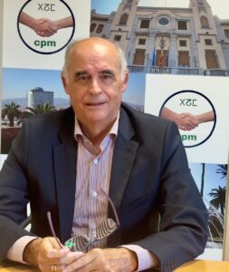 Portrait of an older man in a dark blazer and striped shirt seated at a desk, holding glasses, with a backdrop displaying XZE CPM logos and a building image behind him.
