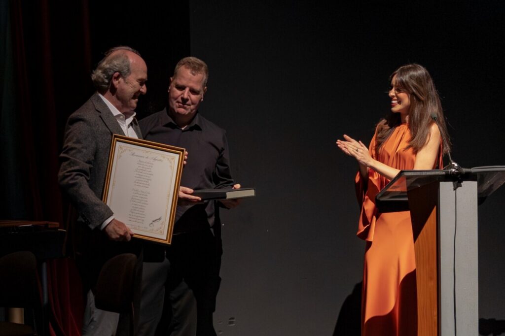 Two men on a stage exchange an award—one holds a large framed certificate while the other hands over a smaller item, as a woman in an orange dress applauds at a podium on the right.
