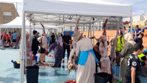 People under white canopies processing animal carcasses during a communal slaughter event; exposed meat, blood on the ground, and volunteers assisting.