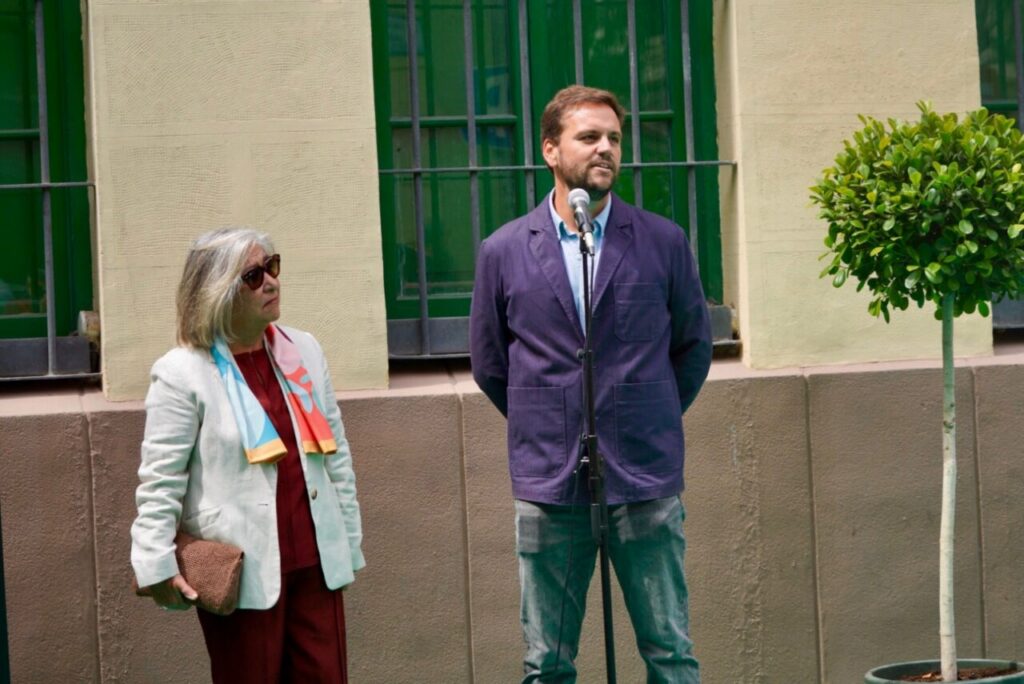 Man in a purple jacket speaks into a standing microphone while a woman in a light blazer and sunglasses stands nearby outdoors. behind them: green windows and a small potted tree.