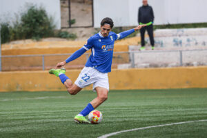 Soccer player in a blue jersey (number 22) kicks a ball on a green field with yellow barriers in the background.