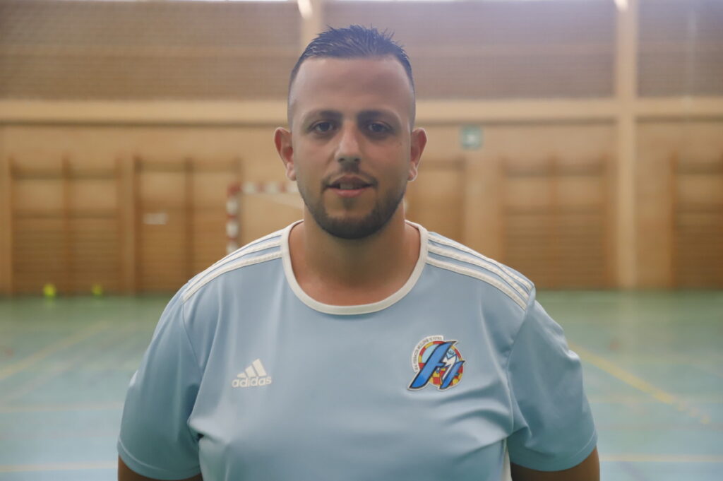 Man in a light blue Adidas sports jersey standing in an indoor gymnasium.