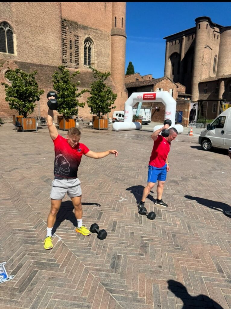 Dos deportistas levantando pesas en un evento deportivo al aire libre