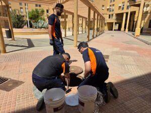 Three workers in navy and orange uniforms repair a manhole, kneeling with tools beside open cover in a sunny courtyard.