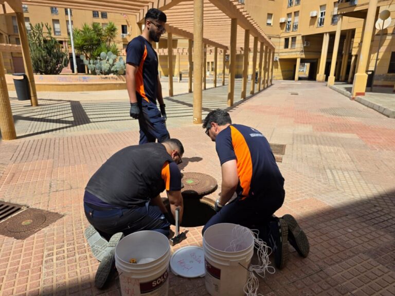 Three workers in navy and orange uniforms repair a manhole, kneeling with tools beside open cover in a sunny courtyard.