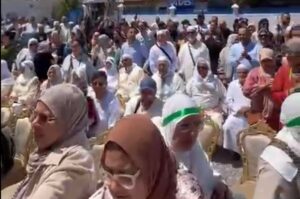 Crowd of people in light clothing seated outdoors at a ceremony, many wearing head coverings and sunglasses.
