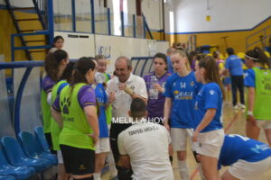 A group of female volleyball players and a coach share a water break on an indoor court, with players in blue jerseys and neon green practice vests gathered around the coach near the bench.
