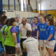 A group of female volleyball players and a coach share a water break on an indoor court, with players in blue jerseys and neon green practice vests gathered around the coach near the bench.
