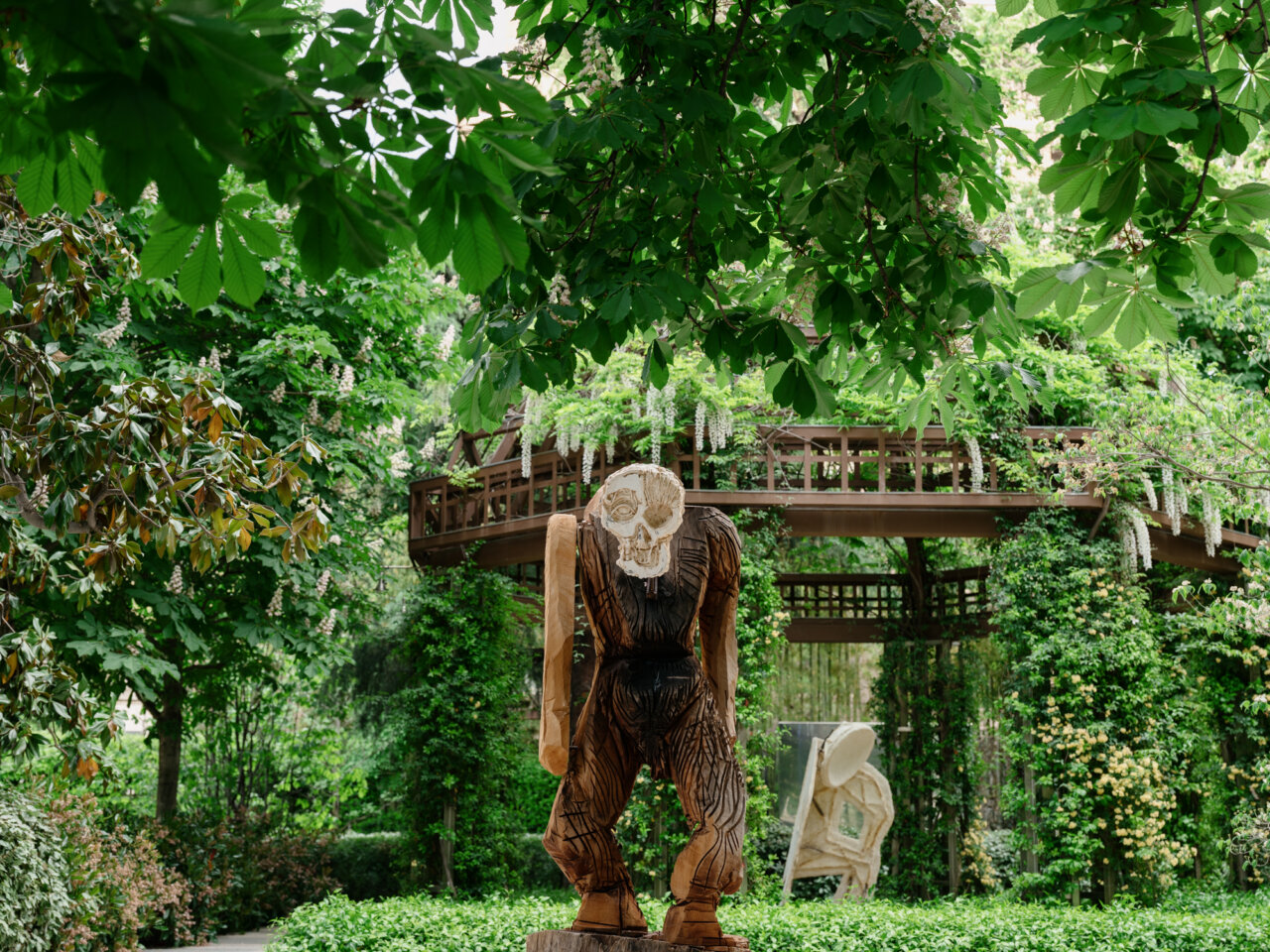 Wooden carved statue of a humanoid figure with a skull head in a lush garden, with a wooden bridge in the background.