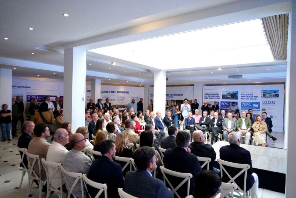Audience seated in a bright conference hall watching a panel on stage with posters in the background on the wall.