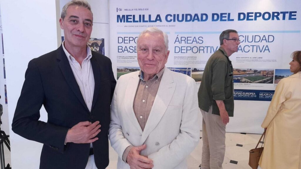 Two men in suits pose for a photo at an exhibition with a large banner that says 'Melilla Ciudad del Deporte' in the background,