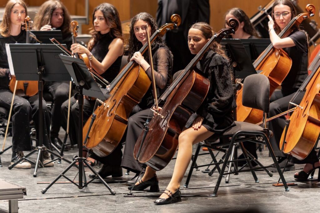 Female cellists in black attire playing cello in a concert orchestra on stage.
