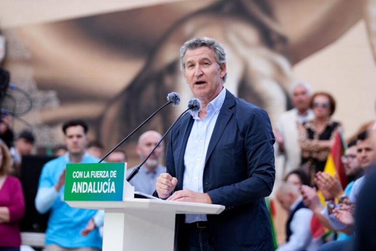 Man in a blue blazer delivers a speech at a white podium with a green sign reading 'Con la fuerza de Andaluceda' during an outdoor rally, microphones in front, crowd clapping behind him.