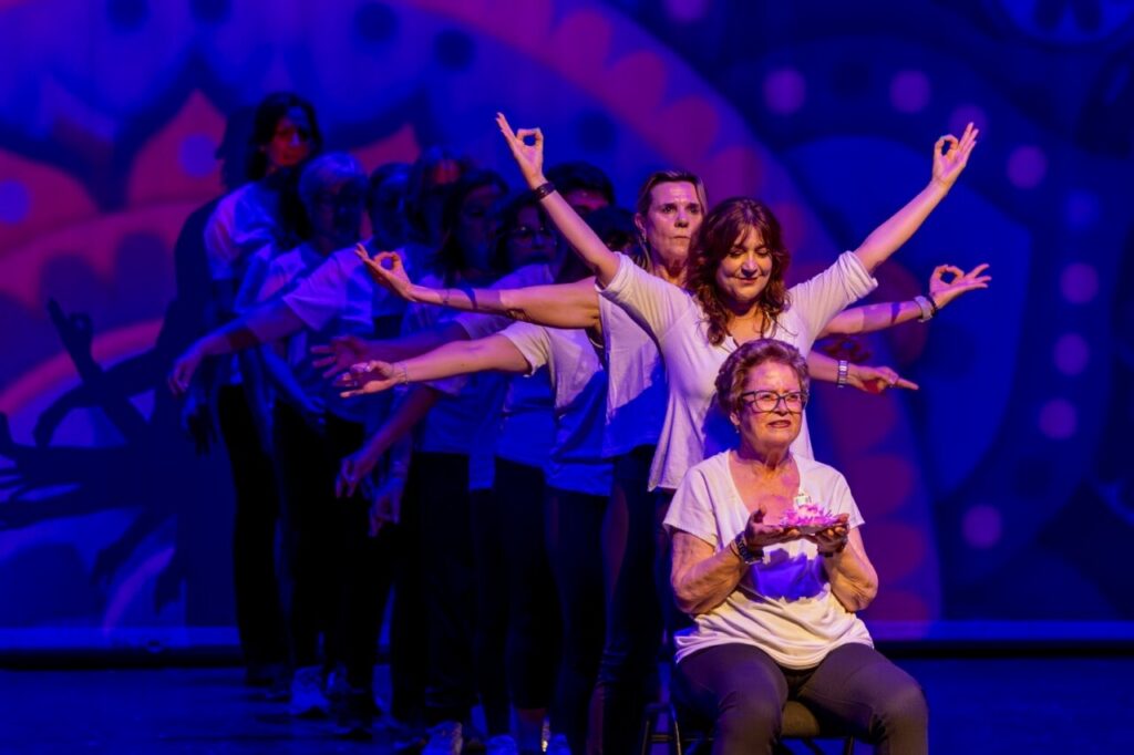 Group of performers on a stage extend their arms in a line; an elderly woman seated in front holds a pink cake under purple-blue lighting, mid-performance.