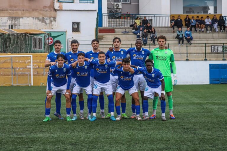 Soccer team in blue uniforms posing for a group photo on a green field with a goalkeeper in a bright green kit and stands in the background.