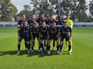 Soccer team group photo: eleven players in navy kits pose together on a green field with a scoreboard in the background.