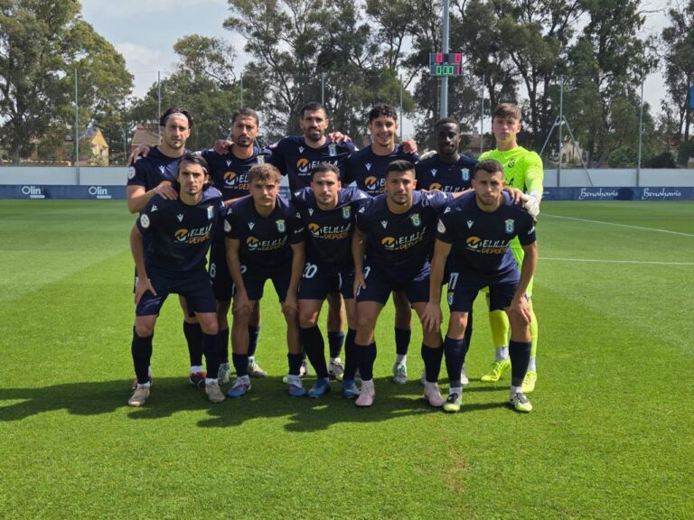 Soccer team group photo: eleven players in navy kits pose together on a green field with a scoreboard in the background.