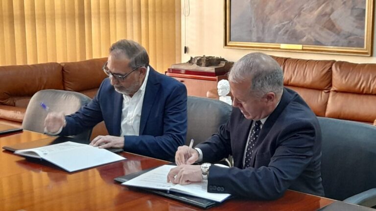 Dos hombres firmando un convenio en una mesa de oficina.