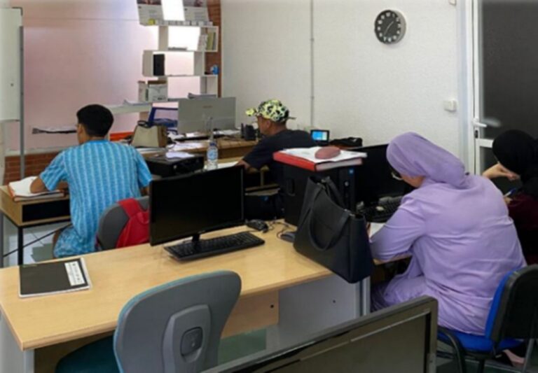 People seated at desks in a computer lab, working on desktop PCs with notebooks and bags nearby.