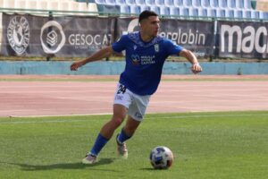 Soccer player in blue jersey dribbles ball on grass field during a match, with banners in the background.
