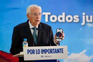 Older man in a suit giving a speech at a podium, holding a phone that shows a pie chart, with a blue backdrop behind him.