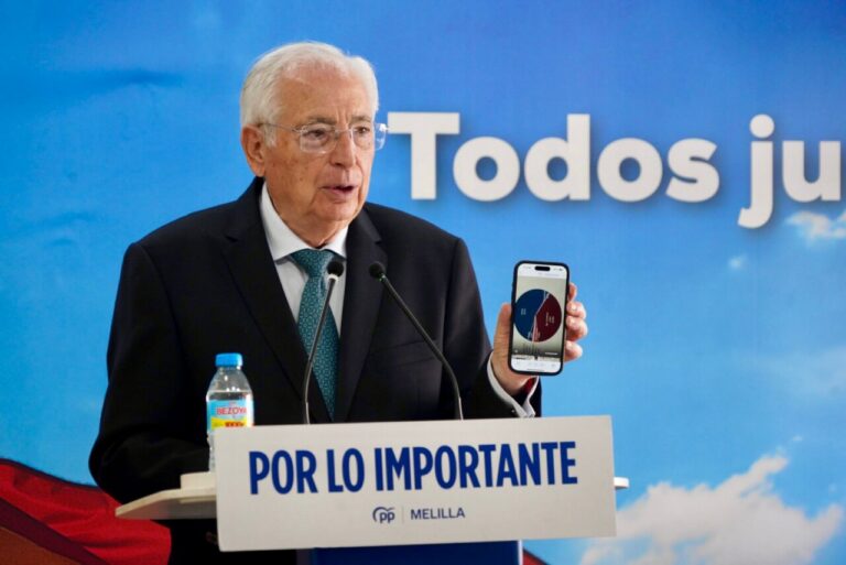 Older man in a suit giving a speech at a podium, holding a phone that shows a pie chart, with a blue backdrop behind him.