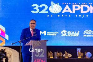 Man in a blue suit speaks at a podium on a stage with a blue backdrop reading '32 APDM' and event logos in the background.