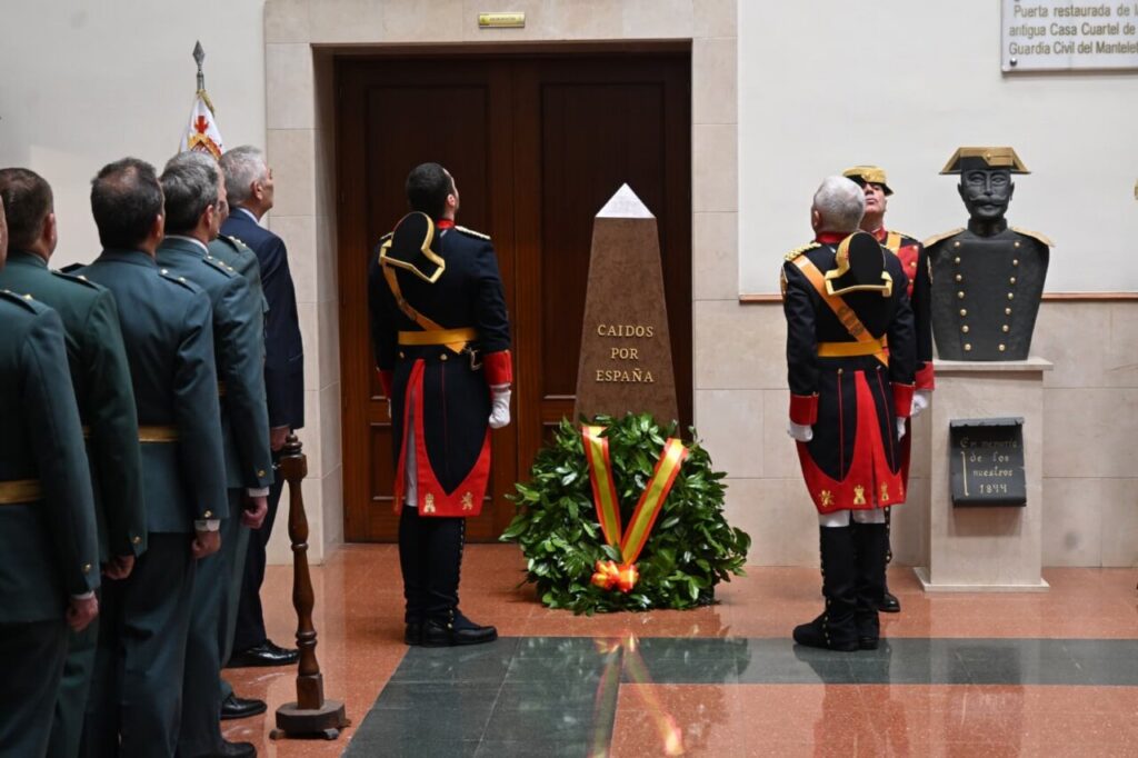 Line of uniformed soldiers standing at attention around a brown obelisk inscribed 'Caídos por España' with a wreath at its base.