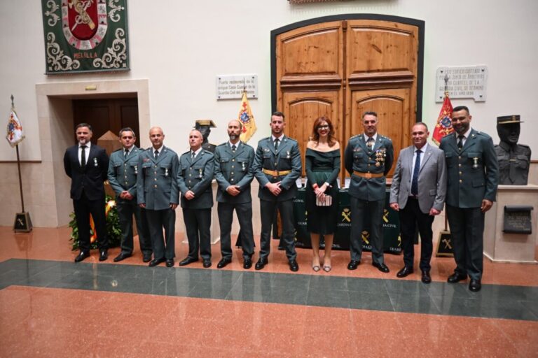 Twelve people, mostly men in green military dress uniforms with medals, stand in a line for a formal ceremony in a hall; a woman in a dark green dress is among them, with wooden double doors and flags behind.