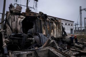 Charred, collapsed industrial equipment at a power facility, with two workers in orange hard hats inspecting the wreckage.