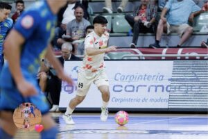 Futsal player in a white kit with orange accents dribbles a pink ball on an indoor court while spectators watch from the stands.