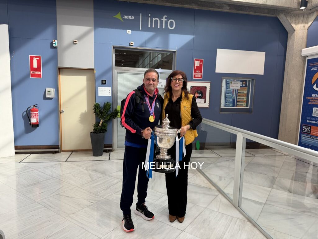Man and woman posing with a large silver trophy adorned with blue ribbons inside a blue-walled info area; both smiling, casual sportswear.