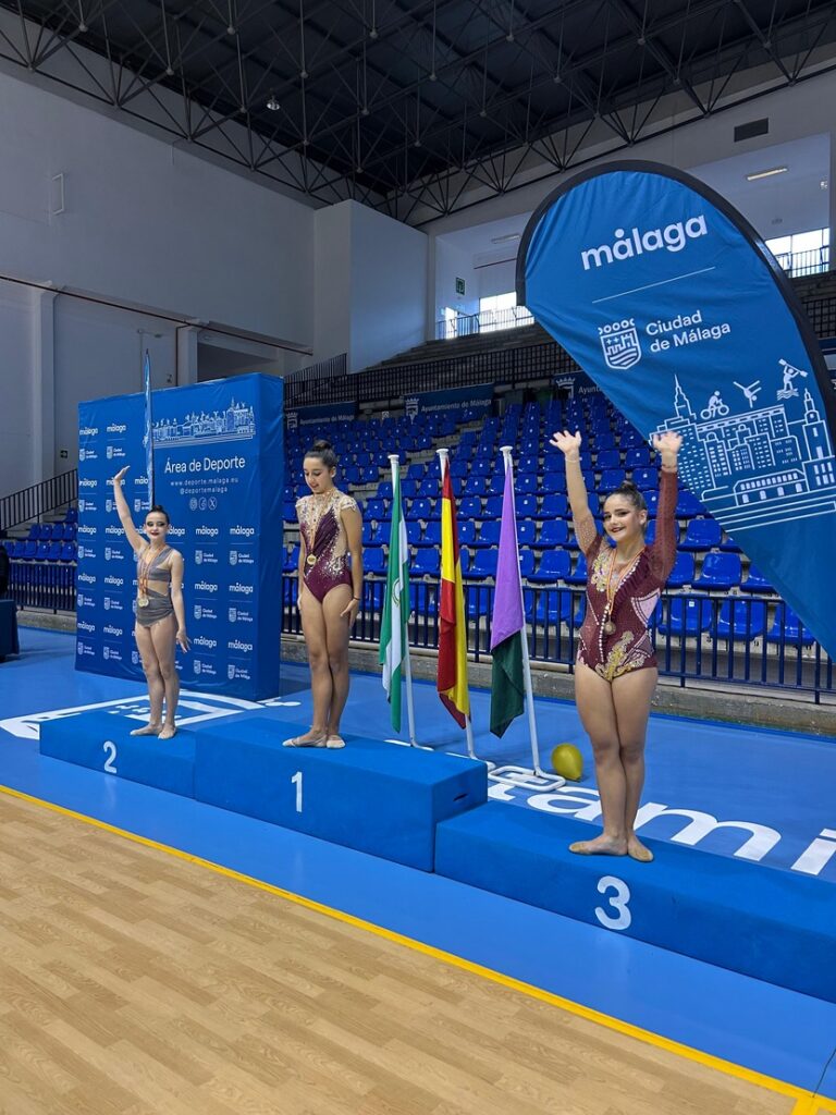 Three young gymnasts stand on a blue awards podium (1st, 2nd, 3rd) during a medals ceremony in a sports hall in Málaga, with blue banners and flags nearby.