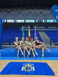 Five young gymnasts in leotards pose on a blue podium in a sports hall, with flags behind and a crest on a mat.