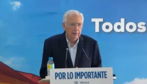 Older man in a dark suit speaks at a podium with a sign reading 'POR LO IMPORTANTE' against a blue backdrop with large white text partially reading 'Todos' behind him.