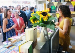 Bustling outdoor book market stall with shelves of books, bunting, and a sunflower bouquet on the counter; a woman in mustard top stands behind the register while a crowd browses nearby.