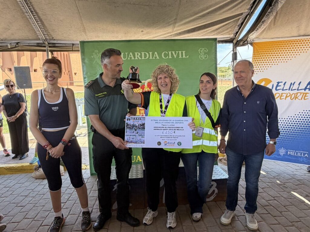 Group of six people on a ceremony stage under a tent; central woman in a neon vest holds a trophy and a large check, flanked by officials and teammates. Green 'Guardia Civil' banner behind them; right banner reads 'Melilla Deporte'.