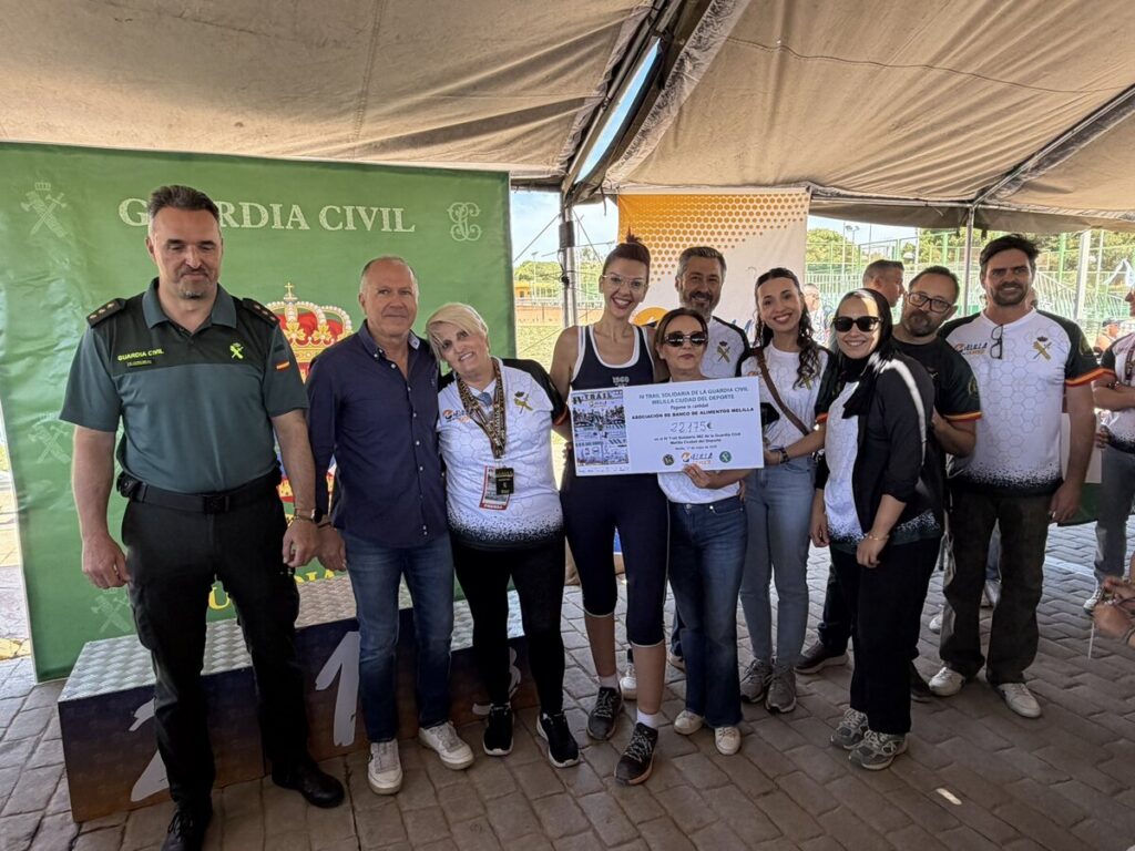 Group of people posing under a tent, including a Guardia Civil officer, with a large ceremonial check and medals on display beside a green backdrop.