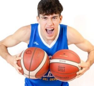 Male basketball player in a blue jersey yelling while holding two basketballs toward the camera.