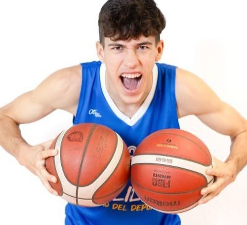 Male basketball player in a blue jersey yelling while holding two basketballs toward the camera.