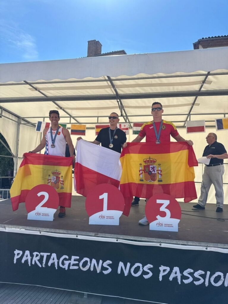 Three male podium finishers with medals on a stage; the center athlete holds the Polish flag, while the left and right athletes display the Spanish flag; banner reads ‘PARTAGEONS NOS PASSIONS’.