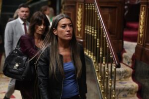 Woman with long hair in a blue top and black leather jacket walks past ornate wooden stairs with gold balusters in a formal interior setting.